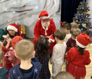 Na zdjęciu widać grupę dzieci zgromadzonych wokół osoby przebranej za Świętego Mikołaja. Mikołaj siedzi na krześle, ubrany w tradycyjny czerwony strój z białym futerkiem i czapkę, a obok niego klęczy kobieta w czerwonej czapce Mikołaja, pomagająca rozdawać prezenty.