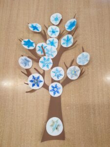 Na zdjęciu widać pracę plastyczną przedstawiającą drzewo zimowe. Pień i gałęzie zostały wykonane z brązowego papieru i przyklejone do jasnego tła. Zamiast liści na gałęziach znajdują się białe kółka z narysowanymi śnieżynkami w różnych odcieniach niebieskiego.

Każda śnieżynka jest inna — niektóre mają delikatne, cienkie ramiona, inne są bardziej geometryczne i wyraziste. Widać, że praca została wykonana przez dzieci, ponieważ rysunki różnią się stylem i poziomem szczegółowości.

Całość tworzy efektowne, zimowe drzewo, które symbolizuje nadejście zimy i zastępuje liście śniegowymi płatkami.
