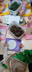 Na zdjęciu widać grupę dzieci siedzących przy stole i wykonujących prace plastyczne o tematyce zimowo‑świątecznej. Na stole leżą kolorowe tacki – żółte, różowe i fioletowe – na których dzieci mają okrągłe kartoniki, które smarują klejem za pomocą pędzelków. Na środku stołu stoją dwa białe koszyki: w jednym znajdują się małe szyszki, a w drugim gałązki sztucznej zieleni przypominające igliwie. Widać też kilka białych, okrągłych elementów, prawdopodobnie ozdób lub fragmentów dekoracji. Dzieci są skupione na pracy, a jedno z nich ma na głowie zieloną ozdobę przypominającą świąteczny stroik lub girlandę. Cała scena ma ciepły, twórczy i świąteczny charakter.