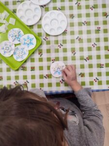 Na zdjęciu widać dziecko siedzące przy stole, które wykonuje pracę plastyczną o zimowej tematyce. Na stole leży cerata w zielono-białą kratkę z motywem czerwonych jagódek. Dziecko trzyma w dłoni okrągły kartonik ozdobiony niebieskimi śnieżynkami, prawdopodobnie tworząc własną ozdobę lub kartkę.

Na stole widać także kilka gotowych prac z podobnym motywem śnieżynek, ułożonych na zielonej tacy, oraz plastikowe palety malarskie z resztkami farby lub kleju. Cała scena przedstawia moment twórczej zabawy plastycznej, pełnej skupienia i zimowego klimatu
