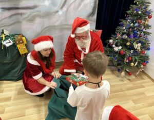 Na zdjęciu widać Świętego Mikołaja i jego pomocnicę wręczających prezent dziecku. Mikołaj ma na sobie tradycyjny czerwony strój z białym futerkiem, czapkę i okulary, a obok niego klęczy kobieta ubrana w czerwono-białą sukienkę i czapkę Mikołaja.

Dziecko, stojące przodem do nich, trzyma w rękach kolorowo zapakowane pudełko z prezentem. W tle po prawej stronie stoi ozdobiona choinka z bombkami, światełkami i łańcuchami, a po lewej widać duży zielony worek z prezentami. 