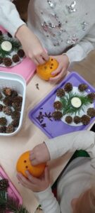 Na zdjęciu widać dzieci wykonujące świąteczne dekoracje. Każde z nich trzyma w dłoniach pomarańczę, w którą wbija goździki, tworząc wzory na skórce owocu. Na stole leżą kolorowe tacki – różowa i fioletowa – z przygotowanymi materiałami do ozdabiania: małymi szyszkami, gałązkami świerkowymi oraz świeczkami typu tealight. W białym koszyku widać więcej szyszek. Dzieci są skupione na swojej pracy, a cała scena ma ciepły, zimowo‑świąteczny klimat, typowy dla zajęć plastycznych w przedszkolu lub szkole.