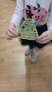 Na zdjęciu widać dziecko trzymające w dłoniach własnoręcznie wykonaną ozdobę w kształcie choinki. Choinka jest zrobiona z zielonego papieru lub tektury, opleciona białym sznurkiem i ozdobiona kolorowymi pomponikami oraz cekinami przypominającymi bombki. Na jej czubku znajduje się duża, błyszcząca gwiazda w kolorze fioletowym. Dziecko ma na sobie białą bluzkę z różowym sercem i motylkami oraz granatową tiulową spódniczkę z drobnymi kropkami. Stoi na drewnianej podłodze, a cała scena ma radosny, świąteczny charakter