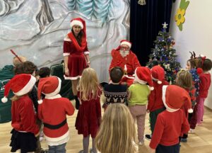 Na zdjęciu widać grupę dzieci stojących w półkolu przed Świętym Mikołajem i jego pomocnicą. Mikołaj siedzi na krześle, ubrany w tradycyjny czerwony strój z białym futerkiem i czapkę, trzymając w rękach prezent. Obok niego stoi kobieta w czerwonej sukience z białymi wykończeniami i czapce Mikołaja, uśmiechnięta i zwrócona w stronę dzieci.