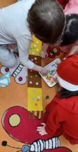 Na zdjęciu widać troje dzieci siedzących na kolorowym dywanie z motywem owadów. Dzieci wspólnie wykonują pracę plastyczną — ozdabiają długi, brązowo-żółty pasek papieru kolorowymi naklejkami w kształcie gwiazdek i kwiatków.

Jedno dziecko ubrane jest w szary strój, drugie w różowy, a trzecie w czerwony kostium z czapką Mikołaja. Na tacy obok nich leżą różnokolorowe ozdoby, które dzieci przyklejają do papieru. Scena ma ciepły, radosny i twórczy charakter, typowy dla zajęć plastycznych w przedszkolu