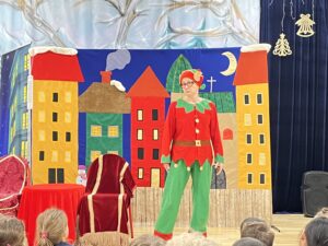 Na zdjęciu widać scenę teatralną przygotowaną dla dzieci. Na środku stoi osoba przebrana za elfa — ma na sobie czerwono-zielony kostium z czapką zakończoną dzwoneczkiem. W tle znajduje się kolorowa dekoracja przedstawiająca bajkowe miasteczko z domkami o czerwonych dachach i żółtych ścianach, a także nocne niebo z księżycem. Na scenie stoją dwa krzesła — jedno czerwone z frędzlami i drugie przykryte czerwonym materiałem. Przed sceną siedzą dzieci, które oglądają przedstawienie. Całość ma świąteczny, radosny klimat.