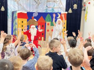 Na zdjęciu widać przedstawienie świąteczne dla dzieci. Na scenie stoją dwie osoby – jedna przebrana za Świętego Mikołaja w czerwonym stroju z białą brodą i czapką, a druga w białym, błyszczącym kostiumie, przypominającym anioła lub postać z zimowej bajki. Za nimi znajduje się kolorowa dekoracja przedstawiająca rząd domków w żywych barwach – żółtym, czerwonym, zielonym i pomarańczowym – na tle niebieskiego nieba z chmurkami.

Przed sceną stoi grupa dzieci, które z entuzjazmem unoszą ręce do góry, uczestnicząc w zabawie lub wspólnej piosence. Wnętrze sali jest jasne i ozdobione zimowymi dekoracjami – na zasłonach wiszą złote dzwoneczki i śnieżne motywy. Atmosfera jest radosna, pełna świątecznej energii i dziecięcego entuzjazmu.
