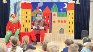 Na zdjęciu widać przedstawienie teatralne dla dzieci. Na scenie siedzą dwie osoby w kolorowych kostiumach — jedna przebrana za elfa w czerwono-zielonym stroju z czapką zakończoną dzwoneczkiem, a druga za starszego mężczyznę z siwą brodą, w niebieskiej czapce i barwnej kamizelce. Za nimi znajduje się kolorowa dekoracja przedstawiająca bajkowe miasteczko z wysokimi, kolorowymi budynkami, zielonym dachem kościoła i nocnym niebem z księżycem. Przed sceną siedzi grupa dzieci, które z zainteresowaniem oglądają występ. Cała scena ma radosny, świąteczny klimat