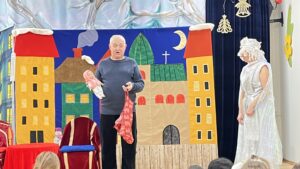 Na zdjęciu widać scenę teatralną przygotowaną dla dzieci. Na środku stoi starszy mężczyzna w szarym swetrze i czarnych spodniach, trzymający w rękach lalkę owiniętą w czerwony materiał. Obok niego, po prawej stronie, stoi osoba przebrana w biały, błyszczący kostium z nakryciem głowy przypominającym śnieżną czapę — wygląda jak postać z bajki, być może anioł lub Królowa Śniegu.

Tło sceny stanowi kolorowa dekoracja przedstawiająca miasteczko z domkami o żółtych i czerwonych dachach, zielonym kościołem z krzyżem oraz nocnym niebem z księżycem. Po lewej stronie widać czerwone krzesło z niebieską poduszką, a przed sceną siedzą dzieci, które oglądają przedstawienie. Całość ma ciepły, świąteczny i bajkowy nastrój.