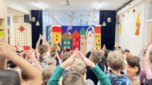 Na zdjęciu widać przedstawienie teatralne dla dzieci odbywające się w przedszkolu lub szkole. Na scenie stoi osoba przebrana w biały kostium, prawdopodobnie odgrywająca postać z bajki lub zimowego spektaklu. Za nią znajduje się kolorowa dekoracja przedstawiająca rząd domków w żywych barwach — czerwonym, żółtym, zielonym i pomarańczowym — na tle błękitnego nieba i srebrnych gałęzi.

Przed sceną siedzi i stoi grupa dzieci, które aktywnie uczestniczą w zabawie — mają uniesione ręce, tworząc z nich kształt przypominający dach domku. Wnętrze sali jest jasne i przytulne, ozdobione świątecznymi dekoracjami, takimi jak girlanda z bombkami i papierowe ozdoby na ścianach. Atmosfera jest radosna i pełna energii.