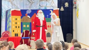 Na zdjęciu widać przedstawienie świąteczne dla dzieci. Na scenie stoją dwie osoby – jedna przebrana za Świętego Mikołaja w czerwonym stroju z białym futerkiem, czapką i długą brodą, a obok niego kobieta w długiej, białej sukni i czepku, przypominająca Panią Mikołajową lub anioła.

Tło sceny stanowi kolorowa dekoracja przedstawiająca rząd domków w żywych barwach – żółtym, czerwonym, zielonym i brązowym – na tle niebieskiego nieba z księżycem i chmurkami. Po lewej stronie stoją dwa krzesła: jedno czerwone z ozdobnym oparciem i drugie niebieskie, a obok nich leży worek z prezentami.

Przed sceną siedzi grupa dzieci, które z zainteresowaniem oglądają występ. Całość ma ciepły, radosny i świąteczny klimat.