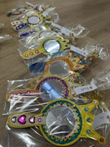 Na zdjęciu widać rząd kolorowych, ozdobnych lusterek zapakowanych w przezroczyste foliowe torebki. Każde lusterko ma dekoracyjną ramkę w kształcie korony lub berła, pomalowaną na żółto, niebiesko, różowo lub pomarańczowo. Ramki są bogato ozdobione błyszczącymi, kolorowymi kamyczkami i naklejkami w kształcie serduszek oraz klejnotów.

Do każdego lusterka dołączona jest mała karteczka z imieniem, co sugeruje, że są to upominki lub rekwizyty przygotowane dla dzieci, prawdopodobnie z okazji balu karnawałowego lub zajęć tematycznych w przedszkolu. Całość leży na drewnianej podłodze, ułożona w równym rzędzie.