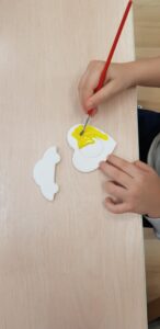 Na zdjęciu widać dziecko malujące pędzelkiem biały element z pianki lub tektury. Kształt przypomina głowę baranka lub owieczki. Dziecko trzyma pędzel z czerwoną rączką i nakłada żółtą farbę na środkową część pracy. Obok, na stole, leży drugi biały element – prawdopodobnie część tej samej figurki.

Stół ma jasne, drewniane wykończenie, a w tle widać fragment podłogi. Scena przedstawia moment zajęć plastycznych, podczas których dziecko maluje elementy dekoracyjne, prawdopodobnie przygotowując magnes na dla dziadków.