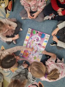 Na zdjęciu widać grupę dzieci siedzących w kręgu na dywanie i grających w kolorową planszową grę edukacyjną. Plansza ma kształt wijącej się ścieżki z polami w różnych kolorach i ilustracjami zwierząt oraz postaci. Dzieci są skupione, każde trzyma pionek lub obserwuje ruch na planszy. Ubrane są w jasne, wygodne ubrania – widać m.in. różowe, szare i beżowe odcienie. Dywan ma wzory w pastelowych kolorach, a cała scena emanuje atmosferą wspólnej zabawy, współpracy i radości z nauki poprzez grę.