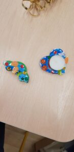 Na zdjęciu widać dwie kolorowe prace plastyczne wykonane z masy plastycznej lub gipsu, leżące na jasnym, drewnianym blacie stołu.

Po lewej stronie znajduje się ozdoba w kształcie skrzydła, pomalowana na zielono i niebiesko, udekorowana kolorowymi guzikami, cekinami i pomarańczowymi elementami przypominającymi listki lub piórka.

Po prawej stronie widoczna jest ozdoba w kształcie serca, pomalowana na niebiesko i ozdobiona pomarańczowymi kwiatkami, zielonymi i czerwonymi guzikami oraz błyszczącymi gwiazdkami. W środku serca umieszczona jest mała świeczka typu tealight.

Całość wygląda jak ręcznie wykonane dekoracje – kolorowe, radosne i starannie ozdobione, prawdopodobnie stworzone przez dzieci podczas zajęć plastycznych.