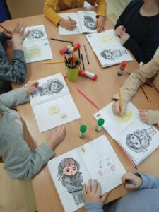 Na zdjęciu widać grupę dzieci siedzących przy stole i zajętych kolorowaniem rysunków w swoich zeszytach. Każde dziecko ma przed sobą kartkę z ilustracją postaci w ubranku przypominającym króliczka oraz rysunkiem okrągłej buzi – prawdopodobnie przedstawiającej emocję. Dzieci używają kredek i ołówków w różnych kolorach, a na stole leżą też kleje w sztyfcie, nożyczki i kubek z przyborami plastycznymi. Uczniowie są skupieni na pracy, a cała scena emanuje spokojną, twórczą atmosferą zajęć plastycznych w przedszkolu lub wczesnoszkolnej klasie.