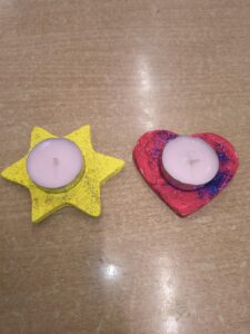 Na zdjęciu widać dwa świeczniki wykonane ręcznie z masy plastycznej lub gipsu. Każdy z nich ma w środku umieszczoną małą świeczkę typu tealight.

Po lewej stronie znajduje się świecznik w kształcie żółtej gwiazdy, ozdobiony delikatnym brokatem, który nadaje mu lekki połysk. Po prawej stronie widoczny jest świecznik w kształcie czerwonego serca, udekorowany niebieskim brokatem, tworzącym efektowny kontrast kolorów.

Oba świeczniki leżą na jasnobrązowym blacie. Całość wygląda jak praca plastyczna wykonana przez dzieci – prosta, kolorowa i radosna, idealna jako ozdoba lub prezent.