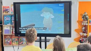 Na zdjęciu widać grupę dzieci siedzących przy stole w przedszkolnej sali i oglądających bajkę na dużym ekranie multimedialnym. Na ekranie wyświetlona jest animowana postać – małe stworzonko o niebieskiej skórze, krótkich włosach i uszach przypominających baranie, ubrane w brązowy sweterek. Na dole ekranu widoczne są napisy dialogowe. Dzieci siedzą tyłem do aparatu, skupione na oglądaniu. W tle widać kolorowe półki z książkami dla dzieci oraz świąteczne dekoracje, w tym małą choinkę i pomarańczową ozdobę w kształcie choinki. Cała scena emanuje ciepłą, edukacyjną atmosferą i zainteresowaniem najmłodszych.