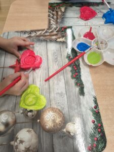 Na zdjęciu widać dziecko malujące farbami gipsowe lub gliniane ozdoby w kształcie serc i gwiazdek. Na stole leży mata z zimowo-świątecznym motywem – widać na niej gałązki świerku, czerwone jagody i bombki w złotych odcieniach.

Dziecko trzyma pędzel i maluje czerwone serce, a obok leży już pomalowane na jasnozielono drugie serduszko. W górnej części zdjęcia znajduje się paleta z farbami w intensywnych kolorach: niebieskim, zielonym i czerwonym. Na macie widać też gotową, niebieską gwiazdkę.

Scena przedstawia moment twórczej pracy plastycznej, prawdopodobnie podczas przygotowywania ozdób z okazji Dnia Babci i Dziadka.