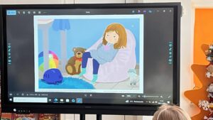 Na zdjęciu widać ekran multimedialny, na którym wyświetlona jest ilustracja przedstawiająca dziewczynkę siedzącą na różowym pufie w dziecięcym pokoju. Dziewczynka ma rude włosy, ubrana jest w niebieską bluzę i granatowe spodnie, a na nogach ma skarpetki w paski. Trzyma w ramionach różowego misia i wygląda na smutną lub zamyśloną. Obok niej siedzi pluszowy miś, kolorowa piłka, niebieski kask i mała figurka zwierzątka. W tle widać niebieski dywanik i fragment baldachimu. Przed ekranem siedzi dziecko, które ogląda ilustrację. Cała scena odbywa się w jasnej, przytulnej sali przedszkolnej z pomarańczowymi dekoracjami i świątecznym drzewkiem po prawej stronie.