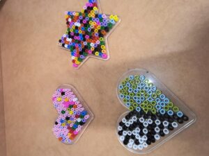 Na zdjęciu widać trzy kolorowe kompozycje wykonane z małych, plastikowych koralików typu Hama Beads, ułożonych na przezroczystych podkładkach w różnych kształtach.

Na górze znajduje się gwiazdka wypełniona wielobarwnymi koralikami – widać odcienie żółtego, różowego, niebieskiego, zielonego, czarnego i pomarańczowego.

Poniżej, po lewej stronie, leży serce w pastelowych kolorach – dominują róż, fiolet, błękit i biel, ułożone w nieregularny wzór.

Po prawej stronie znajduje się drugie serce, wypełnione koralikami w odcieniach zieleni, błękitu, bieli i czerni, tworzącymi geometryczny, kontrastowy wzór.

Całość leży na jasnobrązowym blacie, a kompozycje wyglądają jak prace plastyczne przygotowane do utrwalenia przez zaprasowanie.