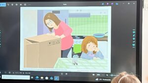 Na zdjęciu widać ekran multimedialny, na którym wyświetlona jest ilustracja przedstawiająca scenę w kuchni. Kobieta w różowej bluzce i okularach pochyla się nad dużym kartonowym pudłem, które stoi na stole przykrytym zielono-białym obrusem w kratkę. Obok niej siedzi dziewczynka o rudych włosach, ubrana w niebieską bluzę, opierająca głowę na dłoni i patrząca z lekkim znużeniem lub smutkiem. Przed nią stoi mała figurka lub zabawka przypominająca szarego kotka. W tle widać kuchenkę, patelnię i zielone płytki na ścianie. Przed ekranem siedzi dziecko, które ogląda ilustrację. Scena ma spokojny, domowy charakter i przedstawia codzienną sytuację w rodzinnej kuchni.