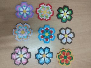 Na zdjęciu widać dziewięć kolorowych kwiatków wykonanych z małych, plastikowych koralików typu Hama Beads. Każdy kwiatek ma inny wzór i zestaw kolorów, ale wszystkie mają podobny kształt – sześć płatków i okrągły środek.

W górnym rzędzie znajdują się kwiatki w odcieniach: fioletowo-niebieskim, czerwono-żółtym oraz zielono-brązowym.
W środkowym rzędzie widać kompozycje w kolorach: pomarańczowo-niebieskim, niebiesko-czerwonym i biało-brązowym.
W dolnym rzędzie znajdują się kwiatki w barwach: różowo-brązowej, niebiesko-czarnej oraz czerwono-zielono-żółtej.

Wszystkie prace ułożone są równym rzędem na jasnobrązowym, drewnianym blacie. Całość wygląda jak efekt zajęć plastycznych, podczas których dzieci tworzyły kolorowe wzory z koralików.
