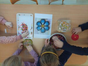 Na zdjęciu widać grupę dzieci siedzących przy stole i układających kolorowe koraliki typu Hama Beads. Na blacie leży kilka pojemników i misek wypełnionych drobnymi, plastikowymi koralikami w różnych kolorach – różowych, żółtych, zielonych, niebieskich, pomarańczowych i białych.

Na środku stołu znajduje się kartka z wydrukowanym wzorem kwiatka z niebieskich i brązowych koralików, który prawdopodobnie służy dzieciom jako przykład do odtworzenia. Obok leży tacka z rozsypanymi koralikami oraz przezroczyste podkładki w kształcie kwiatków, na których dzieci układają swoje wzory.

Dzieci są skupione na pracy, każde z nich wybiera koraliki z pojemników i układa je na podkładkach. Scena przedstawia moment wspólnej, kreatywnej zabawy plastycznej w przedszkolu lub szkole.