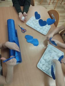 Na zdjęciu widać dzieci siedzące przy stole i wykonujące prace plastyczne. Na blacie leżą dwa styropianowe pojemniki wypełnione białymi kulkami lub kamykami, na których widać narysowane wzory i napisy.

Dzieci trzymają w dłoniach niebieskie, błyszczące serduszka wycięte z brokatowego papieru. Na stole znajduje się również rolka tego samego niebieskiego papieru oraz nożyczki.

Scena przedstawia moment wspólnej pracy plastycznej – dzieci przygotowują ozdoby, prawdopodobnie łącząc białe elementy z niebieskimi serduszkami, tworząc dekorację lub prezent. W tle widać krzesła i fragment sali przedszkolnej.