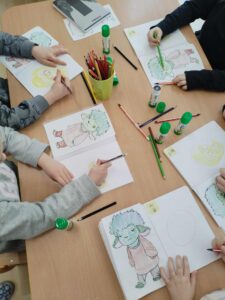 Na zdjęciu widać grupę dzieci siedzących przy stole i kolorujących ilustracje w swoich zeszytach. Każde dziecko ma przed sobą rysunek tej samej postaci – małego, zielonego stworka z dużymi uszami, niebieskimi włosami i ubraniem w odcieniach różu. Dzieci używają kredek ołówkowych w różnych kolorach, głównie zielonych, brązowych i czerwonych. Na stole leżą też kleje w sztyfcie z zielonymi nakrętkami, temperówki, ołówki i kubek z przyborami plastycznymi. Scena przedstawia spokojną, twórczą atmosferę zajęć plastycznych, w której dzieci skupione są na kolorowaniu i rozwijaniu swoich umiejętności manualnych.