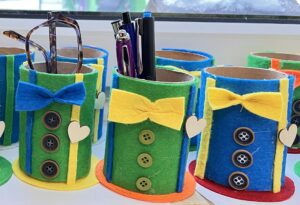 Na zdjęciu widać kolorowe pojemniki na przybory wykonane z rolek po papierze toaletowym, ozdobione filcem i guzikami. Każdy pojemnik ma kształt przypominający cylinder i stoi na okrągłej podstawie z kolorowego filcu.

Rolki są obklejone zielonym, niebieskim lub żółtym filcem, a z przodu mają przyklejone guziki w pionowym rzędzie, co nadaje im wygląd miniaturowych fraków. Na górze każdego pojemnika znajduje się filcowa muszka – żółta lub niebieska – oraz małe serduszko z jasnego materiału. W środku znajdują się długopisy, ołówki i okulary. Całość wygląda jak radosna, kreatywna praca plastyczna wykonana przez dzieci, idealna jako ozdoba biurka lub prezent z okazji Dnia Dziadka.