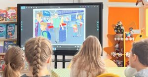 Na zdjęciu widać grupę dzieci siedzących przy stole i oglądających prezentację na dużym ekranie multimedialnym. Na ekranie wyświetlona jest kolorowa ilustracja przedstawiająca ludzi z walizkami na lotnisku. Dzieci siedzą tyłem do aparatu, skupione na oglądaniu. W tle widać jasne, przytulne wnętrze sali przedszkolnej – z pomarańczowymi ścianami, półkami pełnymi książek i ozdobioną choinką, co sugeruje zimowy lub świąteczny czas. Cała scena emanuje ciepłą, edukacyjną atmosferą.