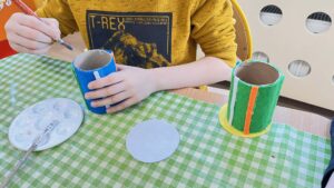 Na zdjęciu widać dziecko siedzące przy stole i wykonujące pracę plastyczną. Na stole leży zielono-biała kraciasta cerata, a dziecko trzyma w ręku pędzelek i maluje lub ozdabia rolkę po papierze toaletowym obklejoną niebieskim filcem. Obok znajduje się druga rolka ozdobiona zielonym filcem z pomarańczowym i żółtym paskiem oraz okrągły kawałek szarego filcu, który prawdopodobnie posłuży jako podstawa pojemnika.

Na stole leży również paleta z białą farbą i pędzelkiem. Dziecko ma na sobie żółtą bluzę z nadrukiem dinozaura i napisem „T-REX”. Scena przedstawia moment twórczej pracy plastycznej, prawdopodobnie podczas zajęć w przedszkolu lub szkole.