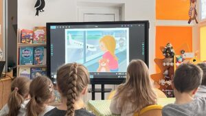 Na zdjęciu widać grupę dzieci siedzących przy stole w przedszkolnej sali i oglądających prezentację na dużym ekranie multimedialnym. Na ekranie wyświetlona jest kolorowa ilustracja przedstawiająca dziewczynkę z misiem w rękach, stojącą przy oknie lotniska, przez które widać samolot. Dzieci siedzą tyłem do aparatu, skupione na oglądaniu. W tle widać jasne, przytulne wnętrze sali – pomarańczowe ściany, półki z książkami i dekoracje świąteczne, w tym małą choinkę. Cała scena tworzy ciepłą, edukacyjną atmosferę pełną ciekawości i zaangażowania.