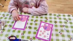 Na zdjęciu widać dziecko siedzące przy stole i wykonujące pracę plastyczną. Na stole leży cerata w zielono-białą kratkę z motywem małych wisienek. Dziecko ma na sobie różową bluzę z nadrukiem postaci z kreskówki i skupia się na ozdabianiu kartki.

Przed nim znajdują się dwie kartki z różową, błyszczącą ramką. Na każdej z nich przyklejony jest różowy słoń z uniesioną trąbą, wykonany z pianki dekoracyjnej. Jedna kartka jest już ozdobiona dodatkowymi elementami – różowymi kwiatkami i fioletowymi serduszkami – a druga jest w trakcie tworzenia. Obok leżą nożyczki i fragment opakowania z ozdobami. Scena przedstawia moment kreatywnej zabawy plastycznej, prawdopodobnie podczas zajęć w przedszkolu lub w domu.