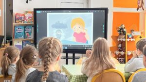 Na zdjęciu widać grupę dzieci siedzących przy stole w przedszkolnej sali i oglądających prezentację na dużym ekranie multimedialnym. Na ekranie wyświetlona jest kolorowa ilustracja przedstawiająca dziewczynkę o rudych włosach w różowej koszulce oraz drugą postać stojącą w deszczu. Dzieci siedzą tyłem do aparatu, skupione na oglądaniu. W tle widać jasne, przytulne wnętrze sali – pomarańczowe ściany, półki z książkami, w tym tytuły dla dzieci, oraz świąteczne dekoracje, takie jak mała choinka i gwiazdy. Cała scena emanuje ciepłą, edukacyjną atmosferą i zaangażowaniem najmłodszych.