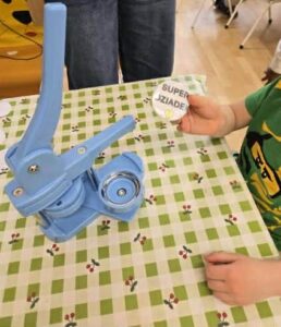 Na zdjęciu widać dziecko siedzące przy stole, na którym stoi niebieska maszyna do robienia przypinek (buttonów). Stół przykryty jest ceratą w zielono-białą kratkę z motywem małych wisienek. Dziecko trzyma w dłoni gotową przypinkę z napisem „SUPER DZIADEK” na białym tle.

W tle widać fragmenty nóg dorosłych osób oraz drewnianą podłogę. Scena przedstawia moment zajęć plastycznych lub warsztatów, podczas których dzieci samodzielnie wykonują przypinki – prawdopodobnie z okazji Dnia Dziadka.