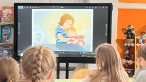 Na zdjęciu widać grupę dzieci siedzących przy stole w przedszkolnej sali i oglądających prezentację na dużym ekranie multimedialnym. Na ekranie wyświetlona jest ilustracja przedstawiająca kobietę w niebiesko-białej bluzce w paski, tulącą małe dziecko z misiem w rękach. Scena na ekranie ma ciepłe, pastelowe barwy, tworzące spokojny i wzruszający nastrój. Dzieci siedzą tyłem do aparatu, skupione na oglądaniu. W tle widać kolorowe wnętrze sali – pomarańczowe ściany, półki z książkami dla dzieci oraz świąteczne dekoracje, w tym małą choinkę i gwiazdki. Całość tworzy przyjazną, edukacyjną atmosferę pełną ciepła i zainteresowania.