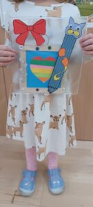 Na zdjęciu widać dziecko trzymające w dłoniach kolorową pracę plastyczną. Praca wykonana jest na przezroczystej, prostokątnej podstawie i ozdobiona różnymi elementami – dużą czerwoną kokardą, niebieskim ołówkiem z główką kota, kolorowym sercem w paski (żółtym, zielonym, pomarańczowym, różowym i niebieskim) oraz małymi błyszczącymi ozdobami w kształcie kwiatków.

Dziecko ma na sobie białą sukienkę w beżowe pieski, różowe rajstopy i niebieskie buty z motywem bajkowym. Stoi na jasnej, drewnianej podłodze, a w tle widać fragment drewnianej ściany. Scena przedstawia moment prezentacji wykonanej pracy plastycznej, prawdopodobnie podczas zajęć w przedszkolu.