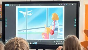 Na zdjęciu widać grupę dzieci siedzących przed dużym ekranem multimedialnym w przedszkolnej sali. Na ekranie wyświetlona jest kolorowa ilustracja przedstawiająca kobietę i dziewczynkę stojące przy oknie lotniska. Dziewczynka trzyma w ramionach pluszowego misia i patrzy w górę na samolot lecący po błękitnym niebie. Kobieta, ubrana w bluzkę w niebiesko-białe paski i z czerwoną torbą na ramieniu, stoi obok niej. Dzieci siedzą tyłem do aparatu, skupione na oglądaniu. W tle widać jasne, przytulne wnętrze sali z pomarańczowymi akcentami. Cała scena emanuje spokojem, ciekawością i atmosferą nauki poprzez zabawę.