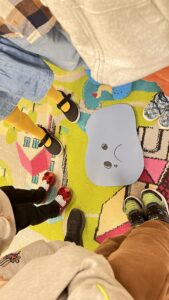 Na zdjęciu widać grupę dzieci stojących w kręgu na kolorowym dywanie w przedszkolu. W środku koła leży wycięta z papieru niebieska chmurka z narysowaną uśmiechniętą buzią. Dzieci mają na sobie różnorodne ubrania i buty – widać m.in. żółte rajstopy, czerwone błyszczące buty, sportowe sneakersy i kapcie. Dywan jest kolorowy, z wzorami przypominającymi domki i ulice. Cała scena wygląda radośnie i przyjaźnie, tworząc atmosferę wspólnej zabawy lub zajęć grupowych.