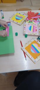 Na zdjęciu widać stół, na którym dzieci wykonują kolorowe prace plastyczne. Na blacie leżą cztery gotowe kartki ozdobione dużym sercem z pasków papieru w różnych kolorach – różowym, niebieskim, żółtym, zielonym i pomarańczowym. Każda kartka ma ramkę z ozdobami: złotymi listkami, błyszczącymi kamyczkami i perełkami.

Na stole znajdują się również przybory plastyczne – kleje w sztyfcie, nożyczki, ołówki, kredki i stos kolorowych pasków papieru. Jedno z dzieci pracuje nad kolejną kartką, przyklejając różowe paski na zielonym tle. W tle widać fragmenty krzeseł i jasną, drewnianą podłogę. Scena przedstawia moment twórczej zabawy plastycznej w przedszkolu lub szkole.