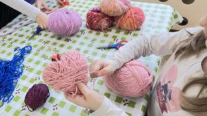 Na zdjęciu widać dzieci wykonujące prace plastyczne z włóczki. Siedzą przy stole przykrytym ceratą w zielono-białą kratkę z motywem wisienek. Jedno z dzieci trzyma w dłoniach różowe serce owinięte włóczką, a w drugiej ręce trzyma kłębek tej samej włóczki.

Na stole leży kilka kolorowych motków – różowych, czerwonych, pomarańczowych i niebieskich – oraz nożyczki. W tle widać drugie dziecko, które również pracuje z włóczką. Scena przedstawia moment kreatywnej zabawy plastycznej, podczas której dzieci tworzą ozdoby z włóczki, prawdopodobnie w przedszkolu lub na zajęciach artystycznych.