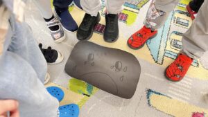 Na zdjęciu widać grupę dzieci stojących w kręgu na kolorowym dywanie. W centrum leży czarna kartka z narysowaną smutną buzią – duże oczy z łzami i opuszczony uśmiech tworzą wyraz smutku. Dzieci mają na sobie różne buty: czerwone z motywem postaci z bajki, niebieskie crocsy, czarne i granatowe sportowe obuwie. Dywan jest jasny, z kolorowymi geometrycznymi wzorami. Scena wygląda na część zajęć przedszkolnych, prawdopodobnie dotyczących emocji lub uczuć.