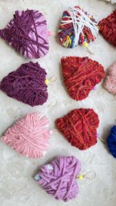 Na zdjęciu widać kilka serduszek wykonanych z włóczki w różnych kolorach. Każde serce ma nieco inny odcień i fakturę – dominują barwy różowe, czerwone, fioletowe oraz jedno wielokolorowe, owinięte dodatkowo białą włóczką.

Serduszka są starannie owinięte grubą przędzą, tworząc miękkie, puszyste ozdoby. Niektóre mają przyczepione małe koraliki lub pętelki ze sznurka, co sugeruje, że mogą służyć jako zawieszki lub dekoracje. Leżą na jasnym, marmurowym blacie, ułożone w nieregularnym układzie. Całość sprawia wrażenie ręcznie wykonanych ozdób, prawdopodobnie przygotowanych przez dzieci podczas zajęć plastycznych.


