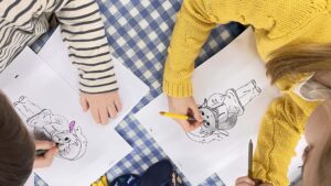 Na zdjęciu widać dwoje dzieci siedzących przy stole przykrytym niebiesko-białym obrusem w kratkę. Dzieci kolorują obrazki przedstawiające postać zwierzątka – prawdopodobnie pieska lub wilczka w ubranku. Jedno dziecko, ubrane w żółty sweter, koloruje rysunek żółtą kredką, a drugie, w bluzce w biało-granatowe paski, używa różowej kredki. Na stole leżą też inne przybory do rysowania. Scena jest jasna, pogodna i pełna skupienia, oddająca atmosferę twórczej zabawy i nauki.