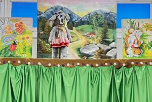 Na zdjęciu widać scenę teatralną, prawdopodobnie przygotowaną dla dzieci. Na środku znajduje się kukiełka przedstawiająca słonika ubranego w różową spódniczkę, szalik i kapelusz. Tło sceny to kolorowy pejzaż górski z drogą prowadzącą przez las i góry. Po bokach znajdują się dwa dodatkowe obrazy – po lewej stronie widać żółwia i biedronkę wśród kwiatów, a po prawej wiewiórkę trzymającą orzech. Dolna część sceny jest ozdobiona zieloną zasłoną z falbaną i brązowym pasem z motywem kwiatków. Całość wygląda bajkowo i radośnie, tworząc atmosferę przedstawienia dla najmłodszych.
