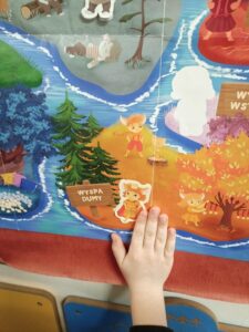 Na zdjęciu widać fragment kolorowej planszy lub plakatu przedstawiającego bajkową mapę. W centralnej części znajduje się obszar oznaczony napisem „Wyspa Dumy”. Na wyspie stoją trzy postacie przypominające małe liski lub inne leśne stworzonka w ubrankach. Wokół nich rosną drzewa — po lewej stronie są zielone świerki, a po prawej drzewa w jesiennych barwach: żółtych i pomarańczowych.

Na dole zdjęcia widać dziecięcą dłoń, która dotyka jednej z postaci na wyspie. W tle widać fragmenty innych wysp oddzielonych niebieskimi rzekami lub kanałami. Całość ma bajkowy, przyjazny i edukacyjny charakter, prawdopodobnie pochodzi z gry planszowej lub materiału dydaktycznego dla dzieci.