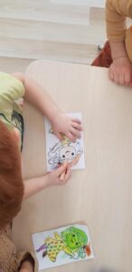 Na zdjęciu widać dwoje dzieci siedzących przy jasnym, drewnianym stoliku. Jedno z nich koloruje obrazek przedstawiający postać czarownicy lecącej na miotle. Używa do tego pomarańczowej kredki. Obok, na stole, leży już pokolorowany rysunek tej samej postaci – ma zieloną twarz, żółtą sukienkę w różowe kropki i pomarańczową miotłę.

Dzieci są skupione na swojej pracy, a scena wygląda na moment zajęć plastycznych w przedszkolu lub w domu. Kolorystyka zdjęcia jest ciepła i jasna, co nadaje mu przyjemny, spokojny nastrój.