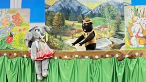 Na zdjęciu widać scenę kukiełkową przedstawiającą bajkowe przedstawienie dla dzieci. Po lewej stronie znajduje się duża kukiełka słonika ubranego w różową spódniczkę, białą bluzkę i kokardkę. Po prawej stronie stoi czarna kukiełka misia w brązowej kamizelce i kapeluszu, który wygląda, jakby rozmawiał ze słonikiem.

Tło sceny to malowany pejzaż górski – widać góry, las, rzekę i drogowskaz z napisem „TATRA”. Po bokach sceny umieszczono kolorowe obrazy z bajkowymi zwierzętami: po lewej biedronkę i żółwia wśród kwiatów, a po prawej białego kota z dużym pomarańczowym ogonem. Dolna część sceny ozdobiona jest zieloną, błyszczącą tkaniną z falbaną i brązowym pasem z kwiatkami. Całość tworzy radosną, kolorową i przyjazną atmosferę teatralnego przedstawienia dla najmłodszych.